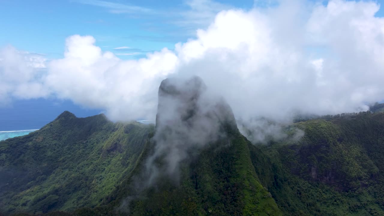 Drone view of a green tropical pacific mountain peak surrounded by clouds on a sunny day in Mouaputa, Moorea, French Polynesia.