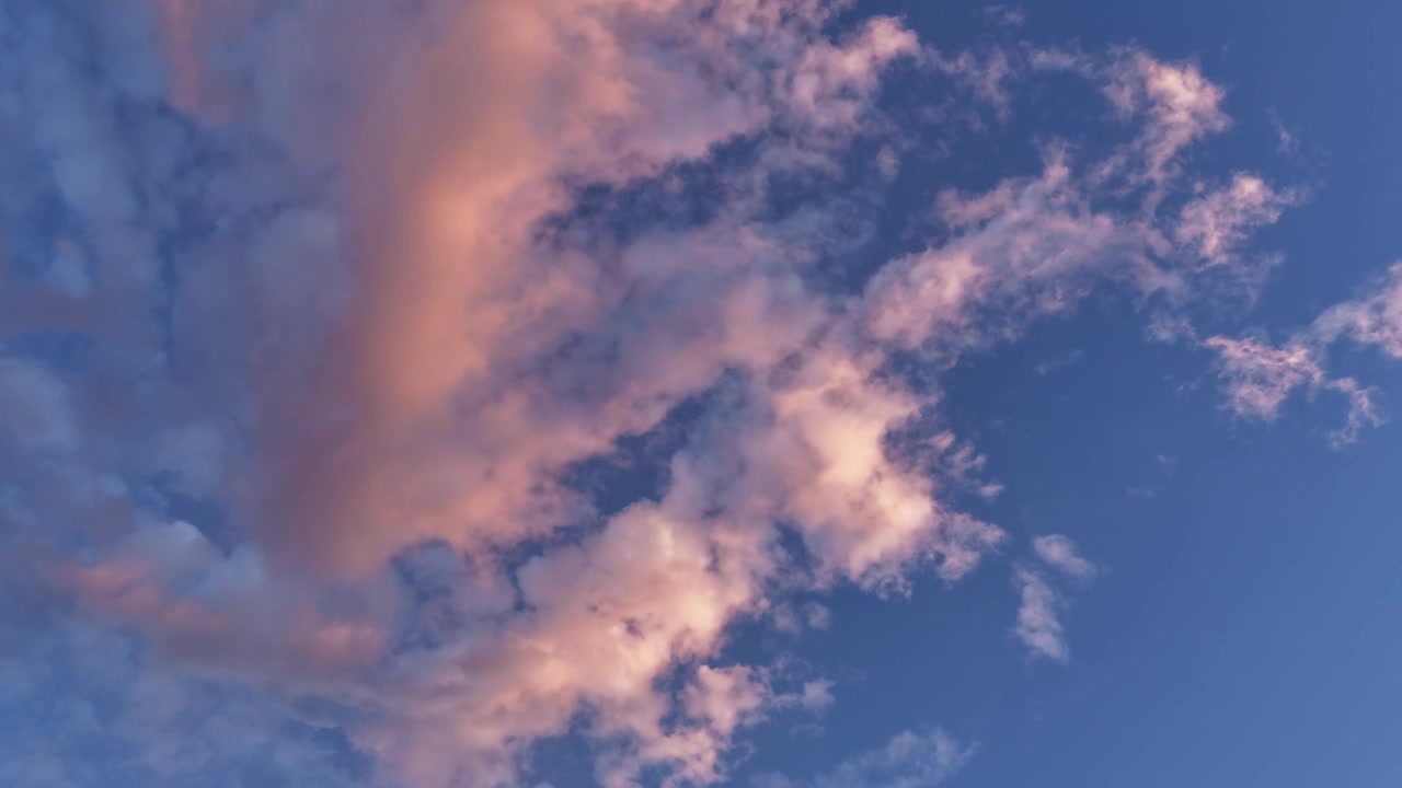 4K UHD timelapse: a textured cloud formation illuminated by warm golden sunset light passes directly overhead. The vibrant peach hues contrast beautifully against the blue evening sky.