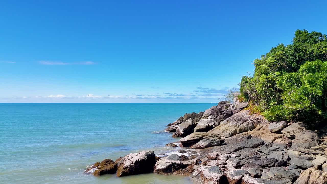 Drone glides above rocky shoreline, turquoise ocean, and lush rainforest under bright daylight skies