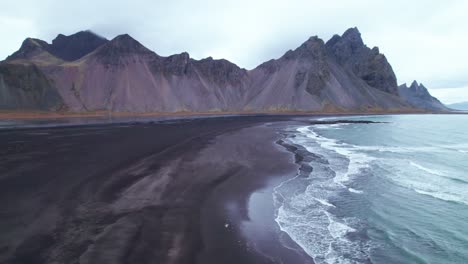 Aérea:-Vestrahorn,-Con-Sus-Icónicos-Picos-Irregulares-Y-Sus-Imponentes-Contrafuertes,-Se-Eleva-Dramáticamente-Desde-Esta-Oscura-Extensión-Como-Una-Colosal-Catedral-De-Piedra.