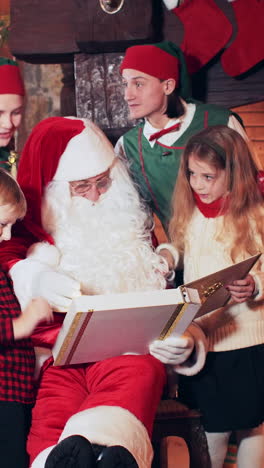santa claus and christmas elves read a book for a boy and a girl near the fireplace with fire and christmas tree with new year decorations. vertical video