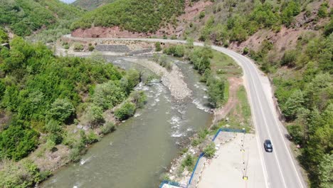 Drone-view-in-Albania-flying-over-a-green-landscape-with-a-rapid-river-and-a-road-next-to-it-with-passing-cars