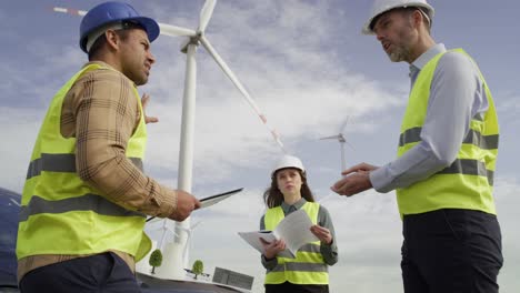 Team-Kaukasischer-Und-Lateinamerikanischer-Ingenieure,-Die-Auf-Dem-Windkraftanlagenfeld-Stehen-Und-über-Das-Projekt-Diskutieren.