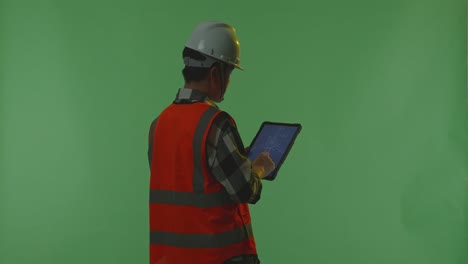 back view of asian male engineer with safety helmet looking at the wind turbine blueprint on a tablet while standing in the green screen background studio, during sunset or sunrise time