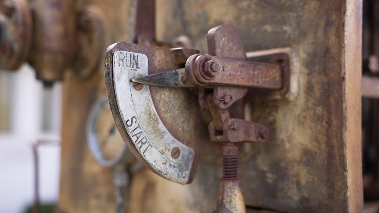 Gammelt industrielt motorkontrolhåndtag, der viser start- og kørepositioner. Rustent mekanisk kontrolhåndtag på en stationær motor, der symboliserer kraft, historie og maskineri.