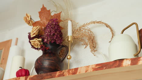 Hand of person retrieves ceramic container from wooden kitchen shelf decorated with autumn-themed floral vase, dry leaves, and rustic accessories, in warm cozy home environment