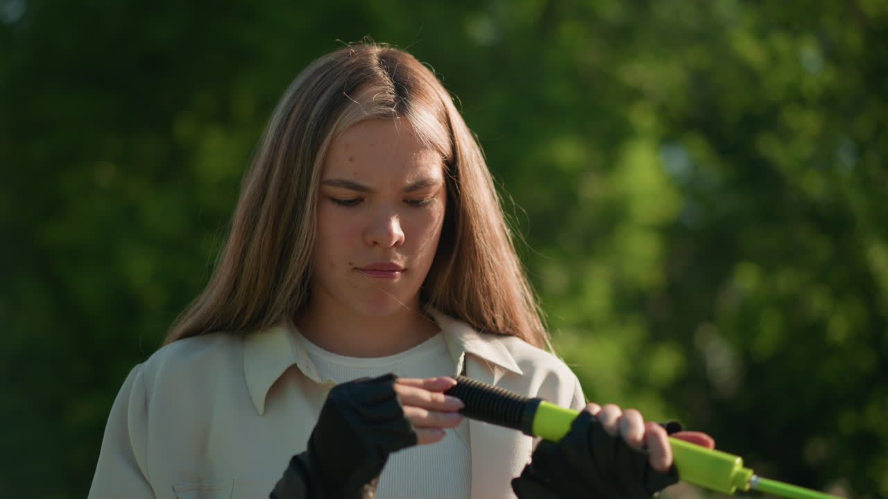 ciclista femenina con cabello rubio inspecciona el mango de la bomba de aire verde con una expresión reflexiva, rodeada por un telón de fondo natural al aire libre con árboles verdes exuberantes