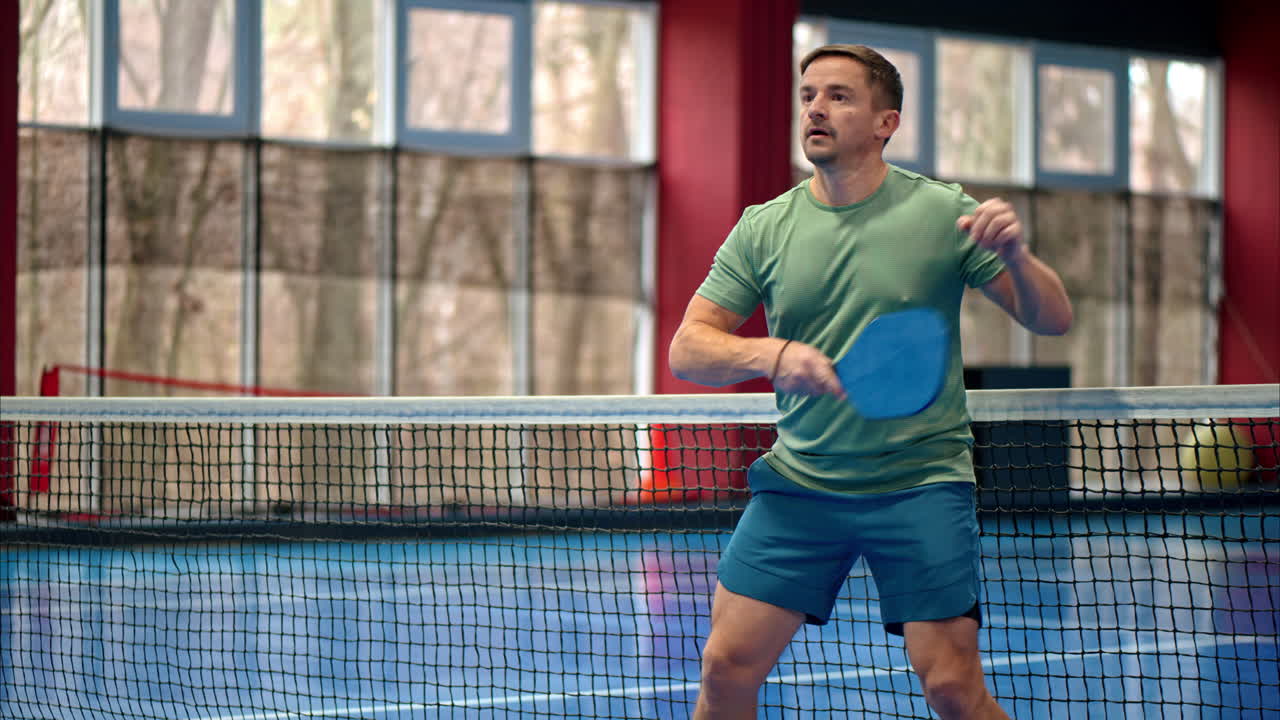 Man in a green shirt playing pickleball on a blue, inside court