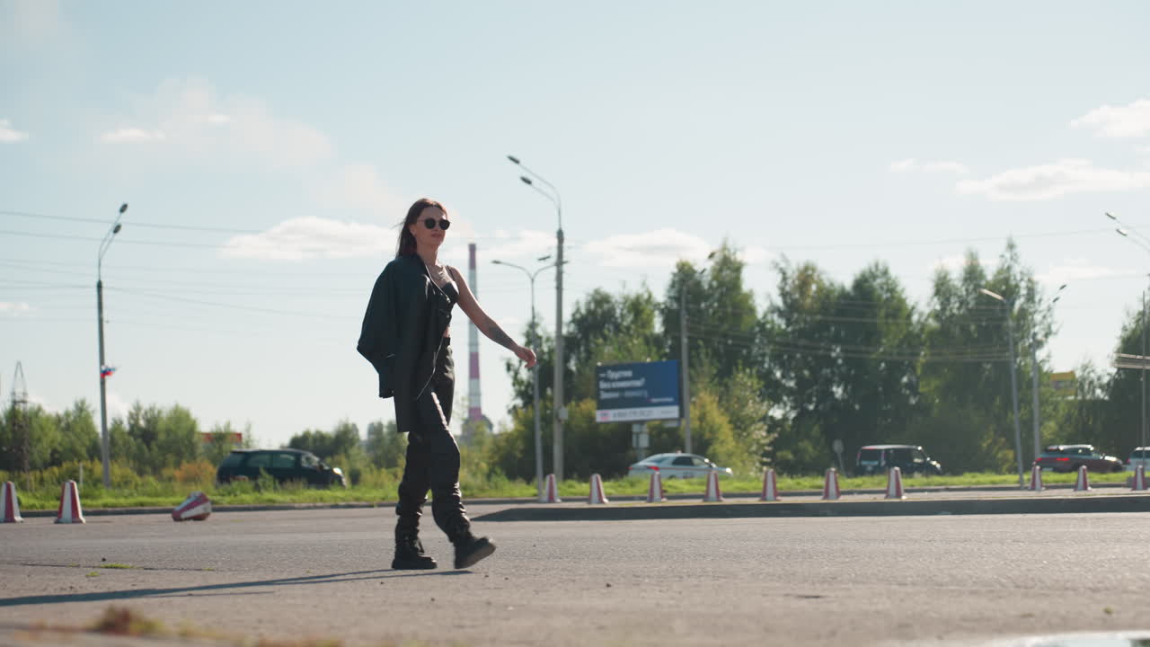 Stylish woman in black outfit walking confidently with jacket over shoulder on city street, sunglasses on, background with passing cars, buildings, power lines, and bright sky symbolizing fashion