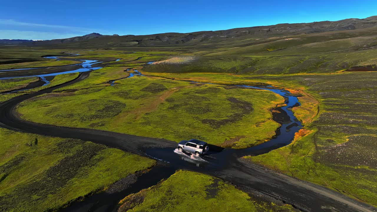 Ripresa con drone di un 4x4 che attraversa un fiume poco profondo su un sentiero di ghiaia attraverso gli altipiani islandesi, circondato da muschio verde e ruscelli tortuosi sotto un cielo estivo azzurro.