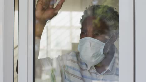 Senior-man-wearing-face-mask-looking-out-of-window-at-home