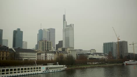 Vista-Del-Día-Nublado-Del-Horizonte-De-Frankfurt-Con-Río-Y-Barcos,-Paisaje-Urbano,-Grúas-De-Construcción-En-Segundo-Plano.