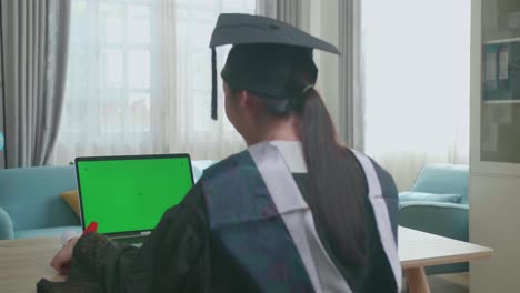 asian woman celebrating on laptop computer with green mock-up screen during an online video call. pretty female graduate wearing a graduation gown and cap sitting on the living room