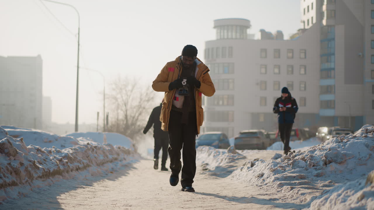 dark skinned man wearing winter coat walking through snow covered street while setting camera and adjusting lens, breathing mist in crisp frosty air amid parked cars and pedestrians near buildings