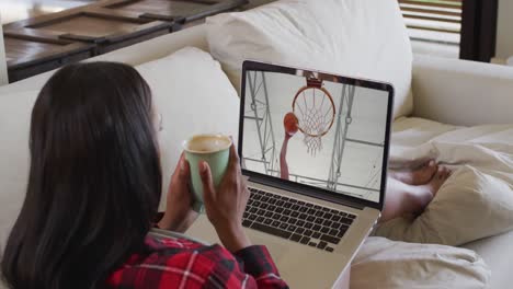 Composite-of-woman-sitting-at-home-holding-coffee-watching-basketball-game-on-laptop