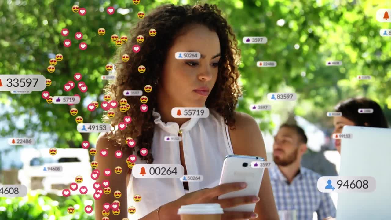 animación de iconos de redes sociales y emojis faciales contra una mujer biracial que usa una computadora portátil en una cafetería