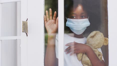 African-american-girl-wearing-face-mask-holding-teddy-bear-waving-through-window