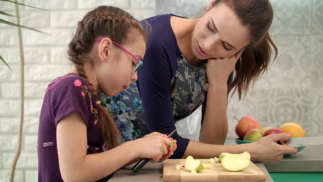 Girl-eating-apple-with-mother-on-kitchen.-Girl-cutting-apple-with-knife