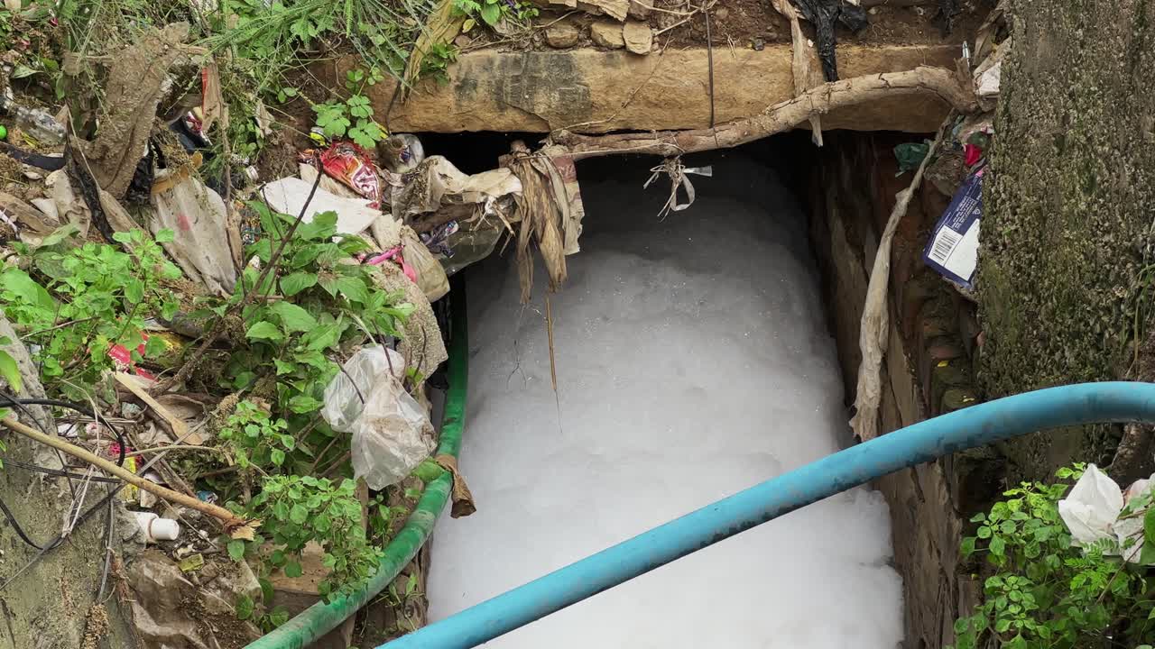 A drainage or water system outlet heavily polluted with a thick layer of white foam, surrounded by significant amounts of trash and debris, indicating a severe environmental issue