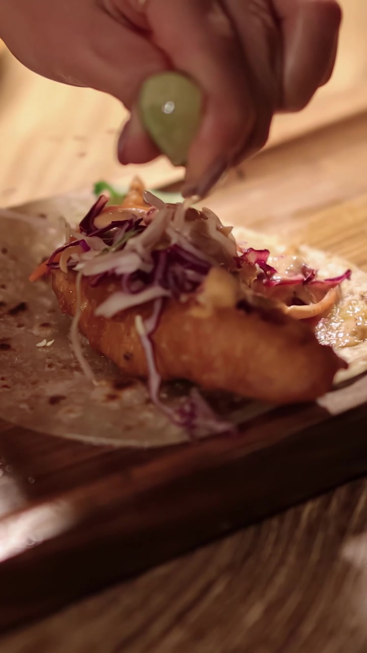 En hånd presser frisk limesaft over en sprød fisketaco toppet med coleslaw og salsa på en blød tortilla, under varm, naturlig belysning.