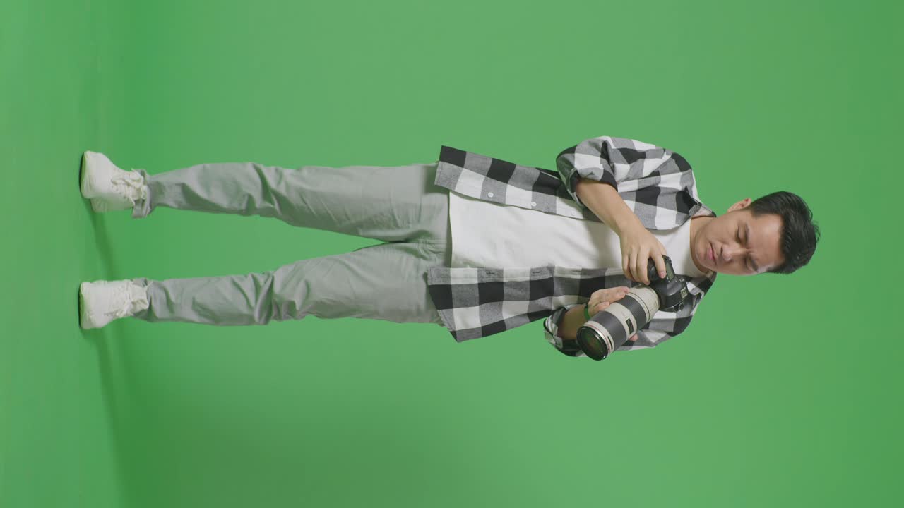Full Body Of Puzzled Asian Photographer Using A Camera Taking Pictures, Saying Why And Making Gestures Doubtfully While Standing On Green Screen Background In The Studio