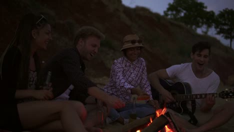 Group-Of-Young-And-Cheerful-People-Sitting-By-The-Fire-On-The-Beach-In-The-Evening,-Grilling-Sausages-And-Playing-Guitar-2