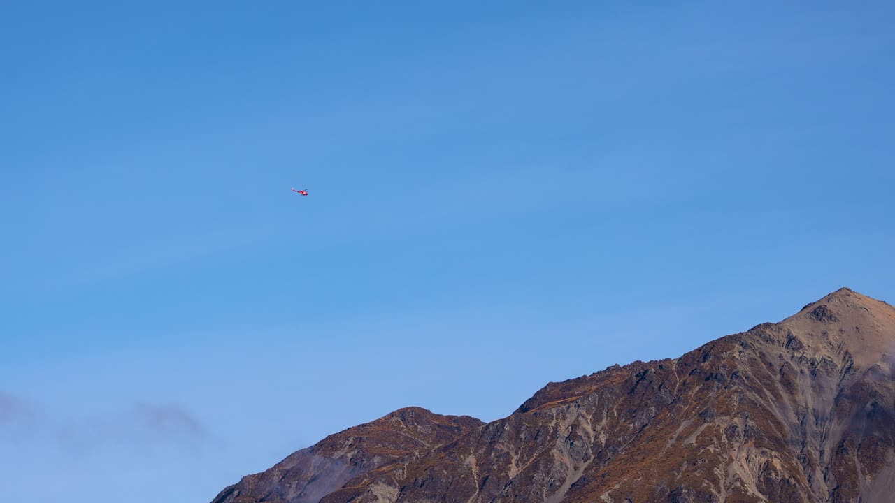 A helicopter flies over a mountain range under clear blue skies in Glenorchy, New Zealand. Captured in natural daylight