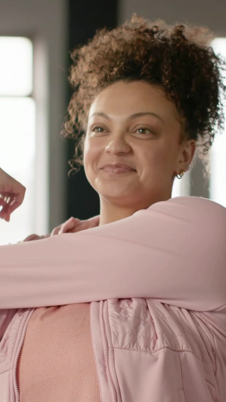 Vertical video: Stretching woman doing chest stretch opening arms in loft by windows in pink jacket