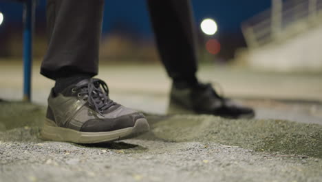 close-up of a person's feet as they repeatedly tap on the ground, with a blurred, dimly lit background and an iron railing visible in the distance