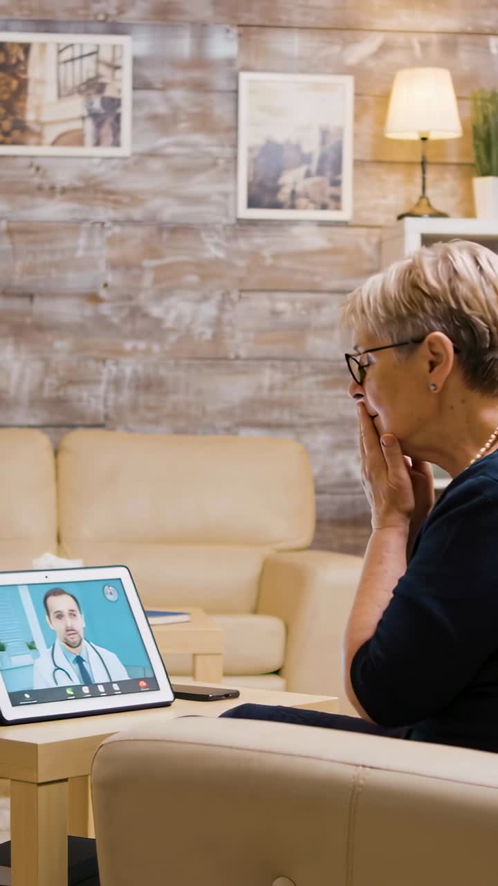 Vertikales Video: Ein Hörakustiker spricht, eine ältere Frau hält die Hand vor den Mund und schaut zu Hause auf ein Tablet.