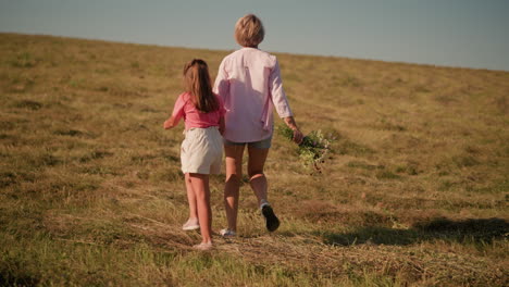 back view of mother and daughter walking together in vast farmland under sunny sky, with mother holding fresh flower bouquet in her right hand, surrounded by vast grass fields