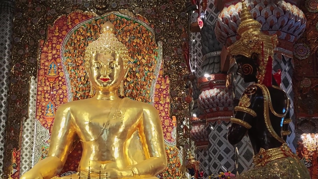 A cinematic rack focus shifts attention from a dark guardian statue with an ornate gold crown to a serene seated golden Buddha statue inside a richly decorated Thai temple. depth-of-field transition
