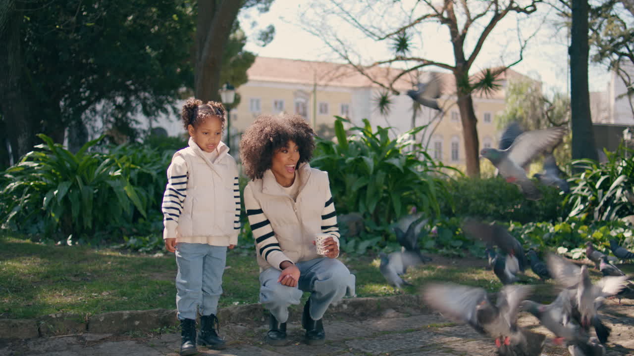 엄마와 함께 푸른 이 있는 공원에서 새를 먹이는 어린 소녀. 머리 위로 날아다니는 비둘기