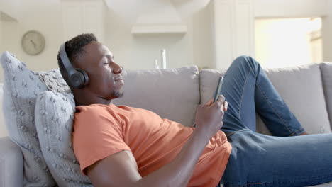 happy african american man using smartphone and headphones in sunny living room, slow motion