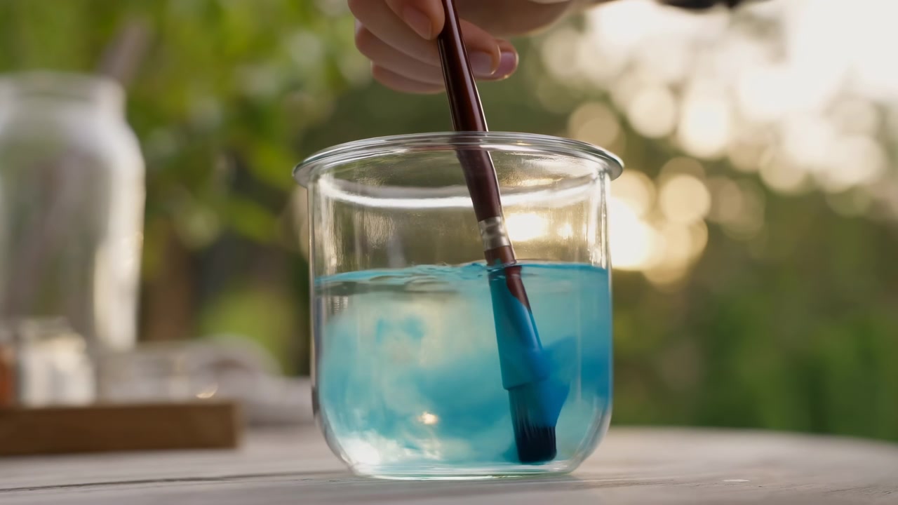 Processo artistico all'aperto che mostra un pennello che si sciacqua in un bicchiere d'acqua a metà sequenza con una calda retroilluminazione
