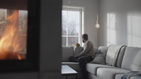 Lonely-Caucasian-Adult-male-holding-tea-cup,-looking-outside-the-window,-Burning-Fireplace-in-Foreground