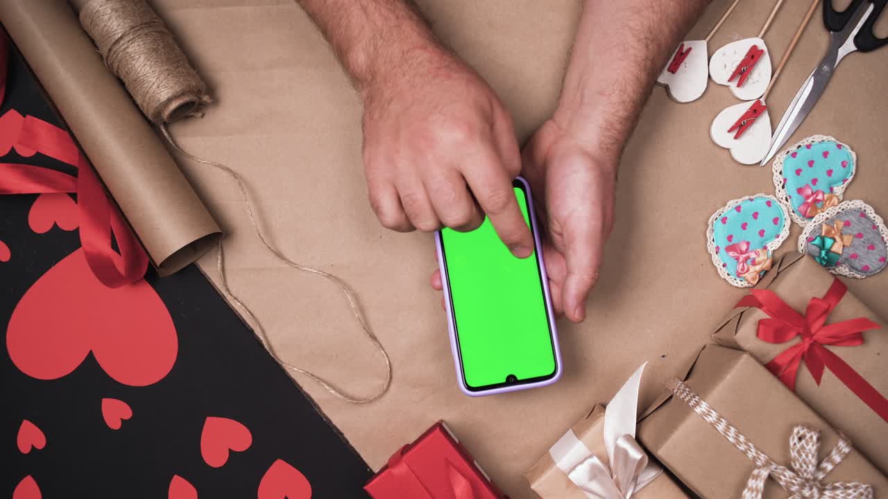 las manos masculinas usan un teléfono inteligente digital con pantalla verde. fondo de mesa con corazones y cajas. concepto de regalo para el día de san valentín