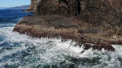 Drone-flies-around-cliff-in-the-ocean,-birds-are-seen-landing-and-standing-on-the-rocks