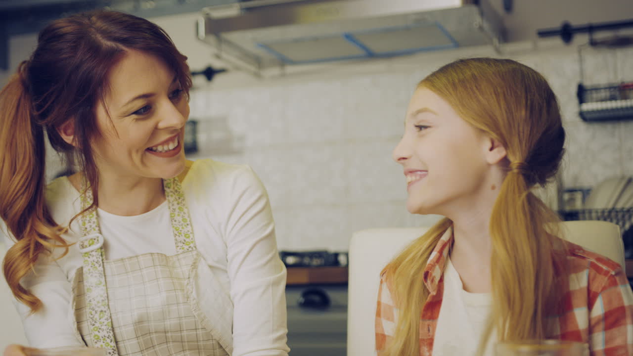 Attractive mother in the apron drinking juice and her cute daughter kissing her in the cheek, than mom and daughter kissing and hugging in the kitchen. Indoors
