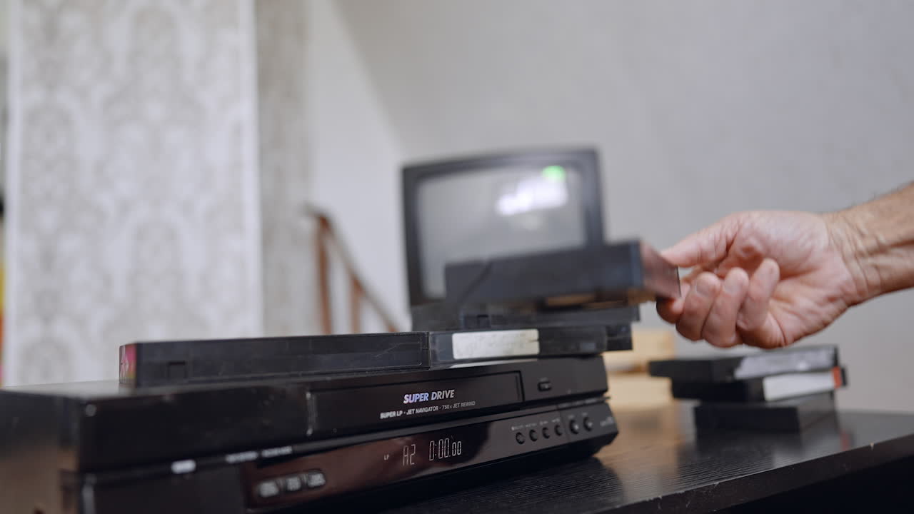 DVD player gives away the tape. Male hand takes the tape. Old tv switched off screen at backdrop