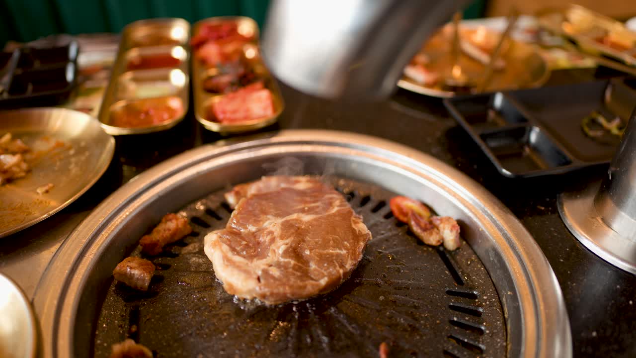 Raw pork steak cooks on tabletop grill with side dishes, warm lighting, and shallow focus