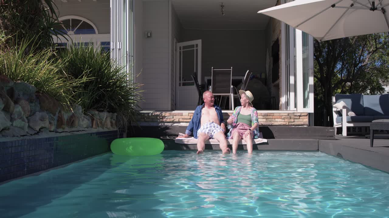 Senior couple sitting pool edge dipping feet, talking, adjusting sun hat, leaning toward green tube