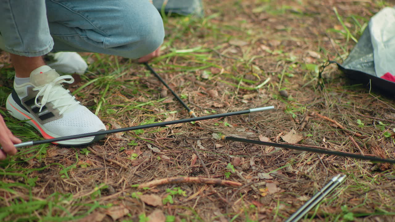 Closeup of couple assembling tent poles on forest floor, hands working together over dried leaves and pine needles, showcasing teamwork and outdoor camping activity in nature setting