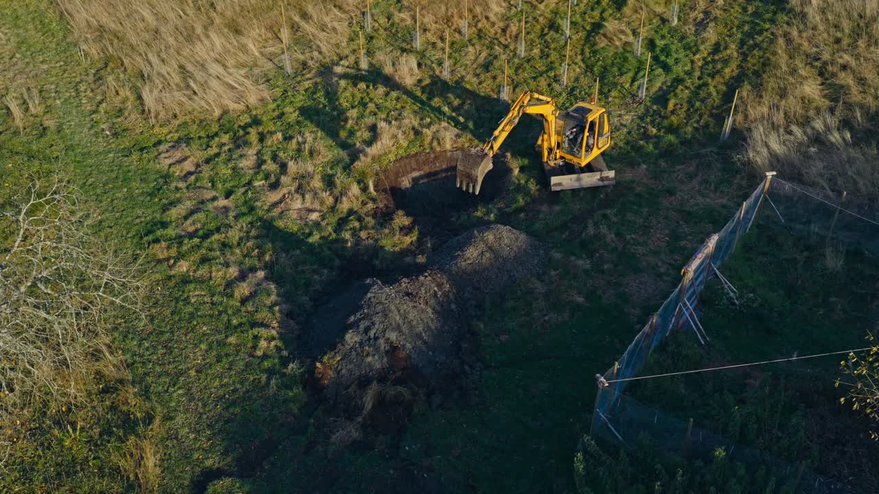 En gravemaskine graver i jorden for at skabe en dam på en græsmark - Orbit droneskud.