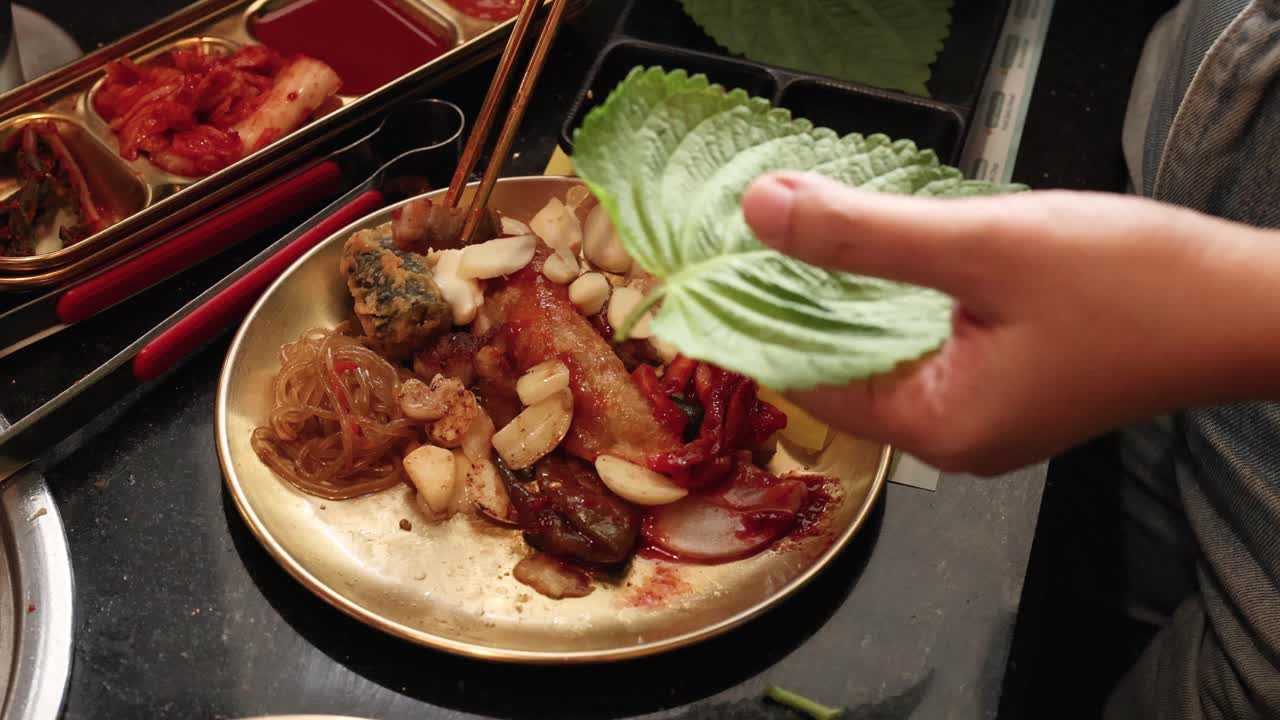 Person assembling Korean BBQ wrap using perilla leaves, grilled meat, and side dishes, overhead view
