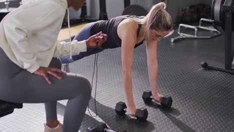 Video-of-diverse-female-fitness-trainer-and-determined-woman-raising-dumbbells-working-out-at-a-gym