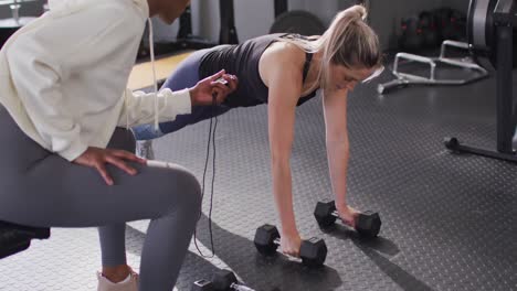 video of diverse female fitness trainer and determined woman raising dumbbells working out at a gym