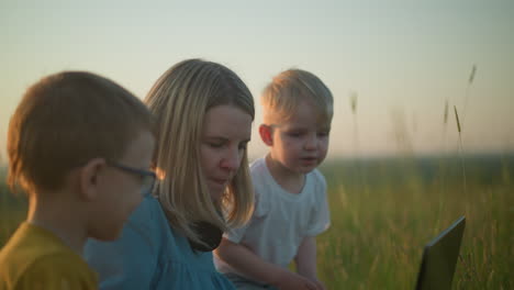 a mother in a blue gown sitting on a grassy field, holding a laptop. her two young sons, one in yellow and the other in white, stand and sit beside her, curiously watching her work