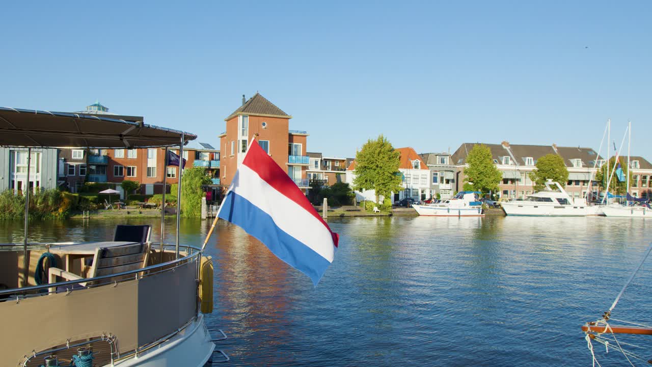 A Dutch flag flutters on a boat in bright daylight, with historic canal-side buildings, calm water, and a bascule bridge in Haarlem, Netherlands