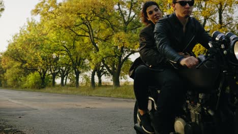 Young-attractive-couple-starting-to-ride-a-motorcycle,-traveling-together-on-the-asphalt-road-on-a-sunny-day-in-forest-in-autumn